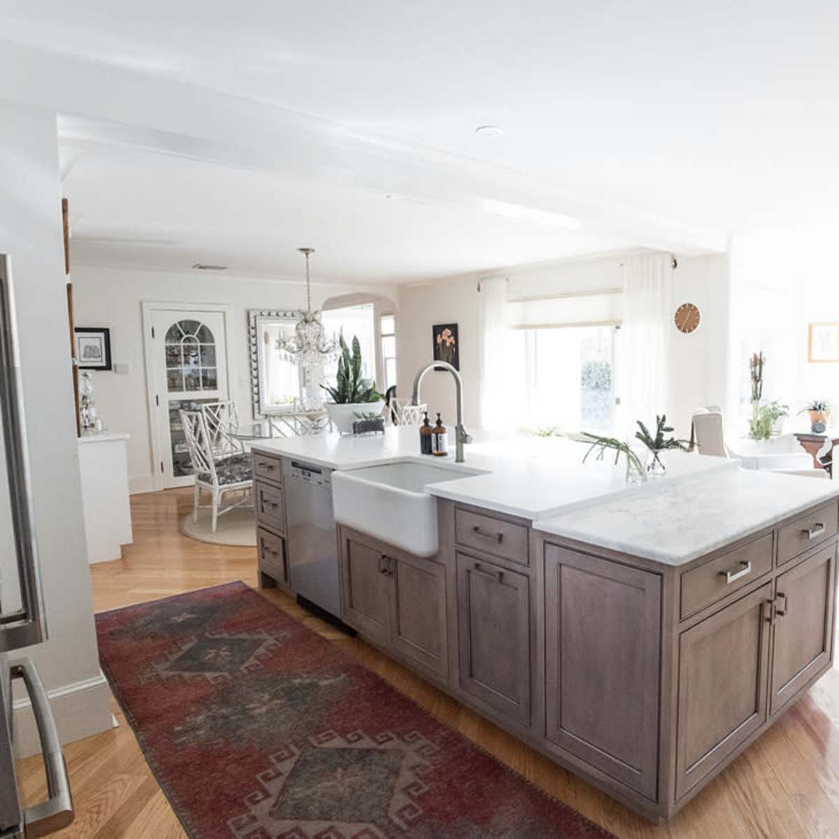 A bright, open kitchen with a large island, featuring a farmhouse sink and modern appliances, adjacent to a living area with windows.