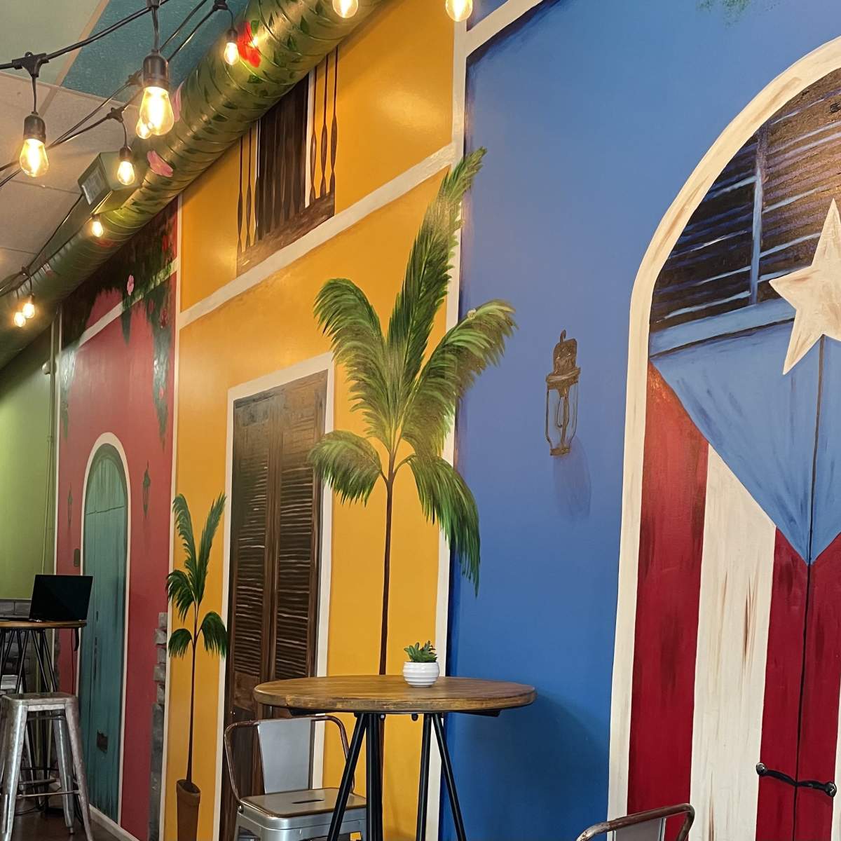 A colorful mural featuring a blue and red door with a white star, surrounded by palm trees and bright walls in a cafe setting.