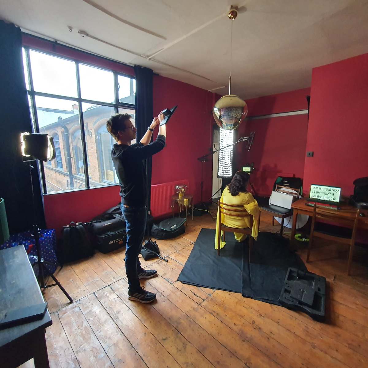 A person is setting up a camera to photograph a model sitting on a chair in a room with red walls and a disco ball hanging from the ceiling.
