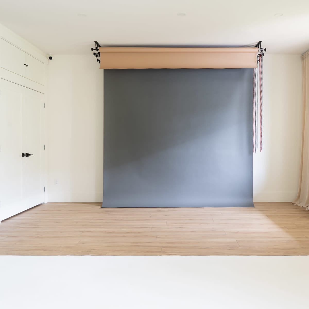 A spacious room with light wooden flooring, a gray backdrop on rollers, and a bench beside a window.