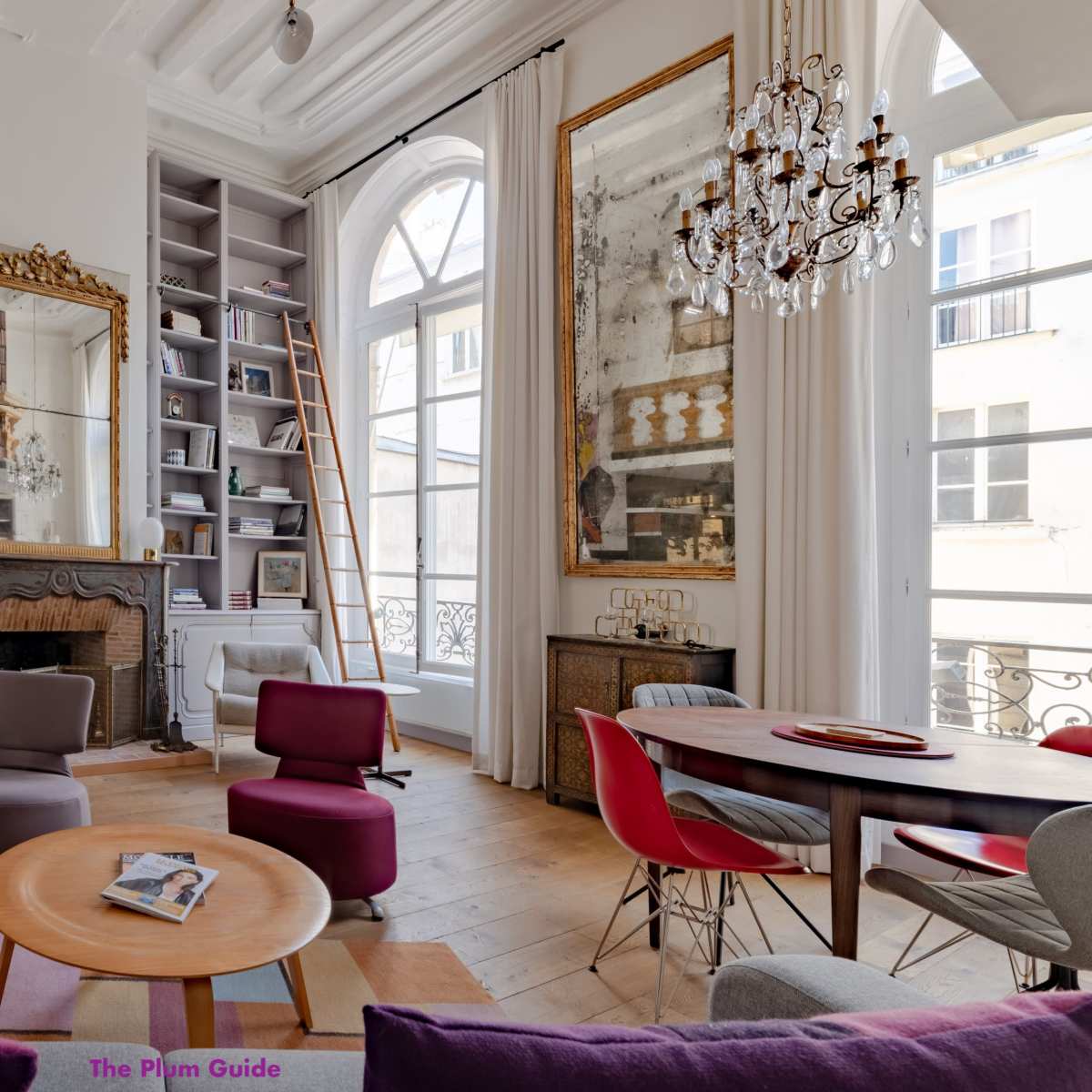 A stylish living room features large windows, a modern art piece, mirrored walls, and a blend of colorful furniture, including a circular coffee table and a dining area.