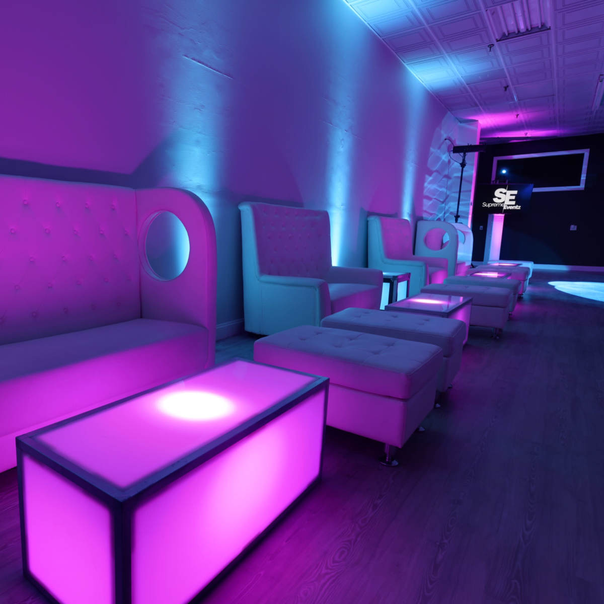 A modern lounge area with purple and blue lighting, featuring stylish couches and illuminated coffee tables.