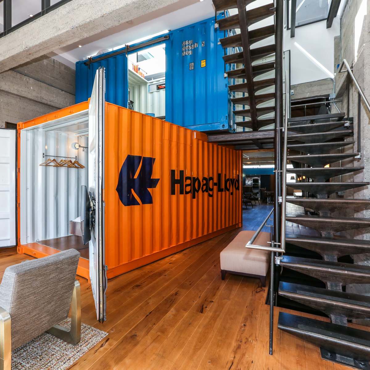 A modern interior space featuring two stacked shipping containers, one orange and one blue, with a staircase leading to an upper level.