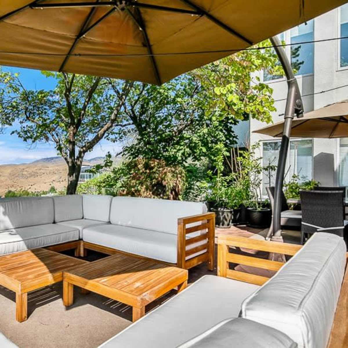 A rectangular outdoor seating area features a sectional couch with light cushions and a wooden coffee table, surrounded by trees and a view of hills in the background.