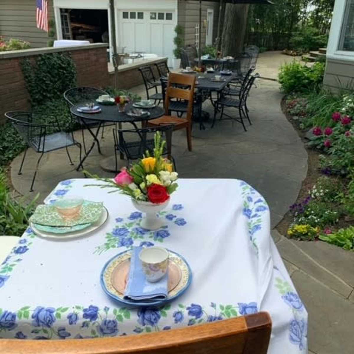 An outdoor dining area features tables set with floral tablecloths and chairs, surrounded by lush greenery and blooming flowers.