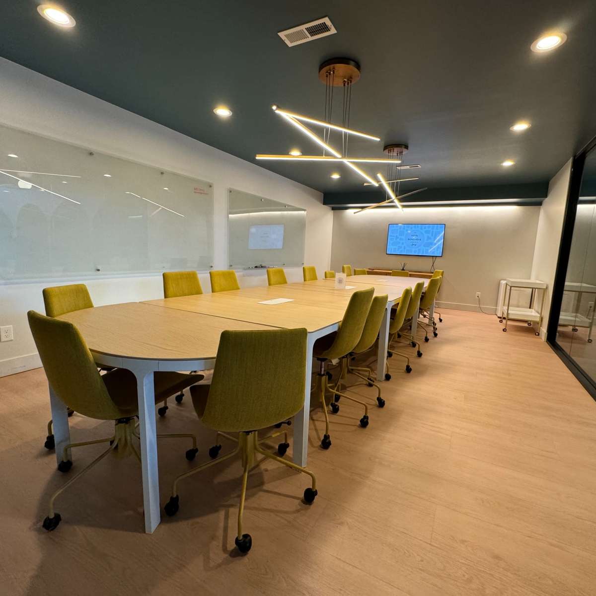 A modern conference room with a long, rectangular table surrounded by chairs and two screens on the walls.