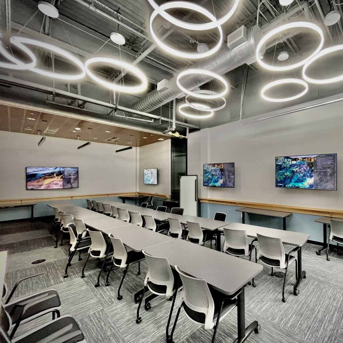 A modern classroom features circular light fixtures and two mounted televisions on the walls, with rows of tables and chairs arranged for instruction.