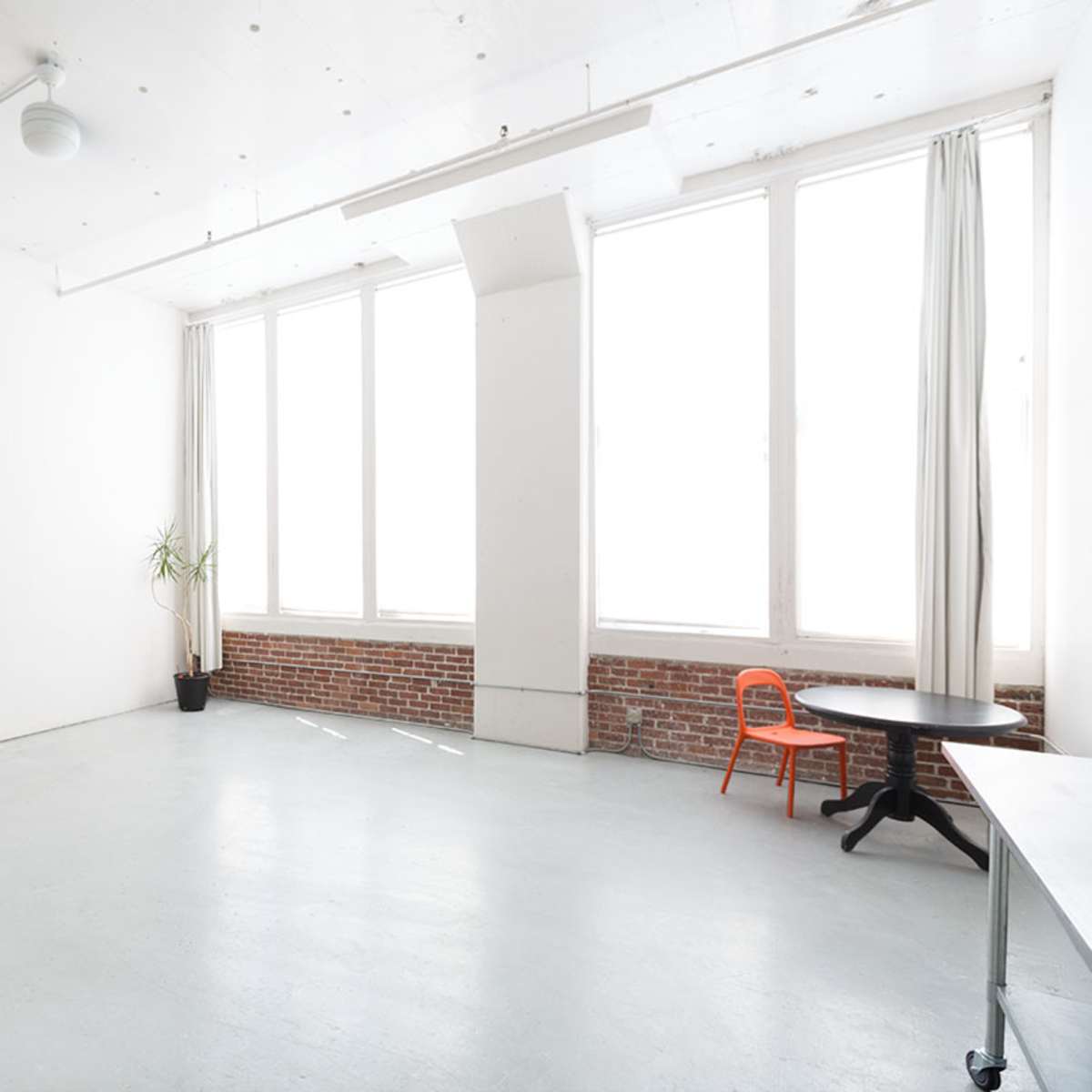 A spacious, empty room with large windows, a brick wall, and minimal furniture including an orange chair and a table.