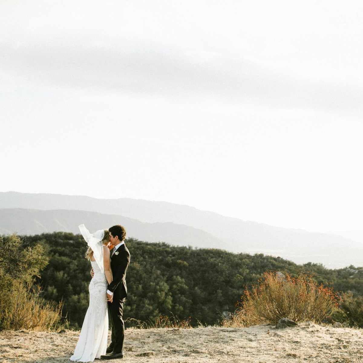 A bride and groom embrace on a hillside with rolling mountains in the background.