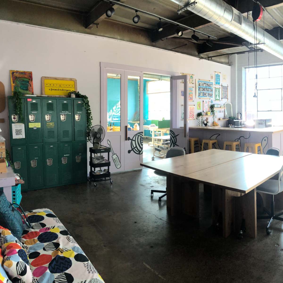 A spacious, brightly lit room featuring a large table, green lockers, a kitchen area, and various workstations.