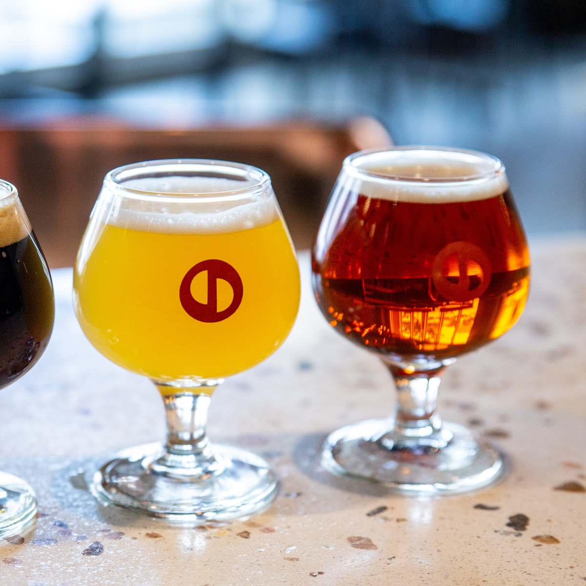 Three distinct glasses of beer are placed on a speckled countertop, featuring a dark brew, a golden lager, and a reddish amber beer, each with a circular logo on the side.