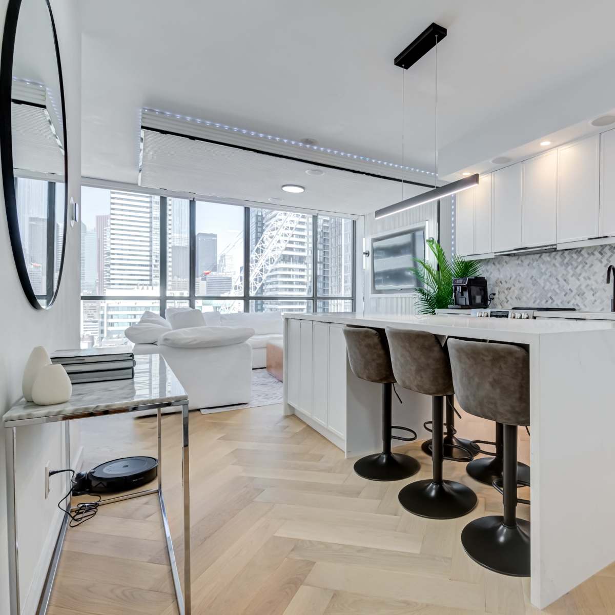 A modern kitchen features white cabinetry and a central island with four bar stools, overlooking a city skyline through large windows.