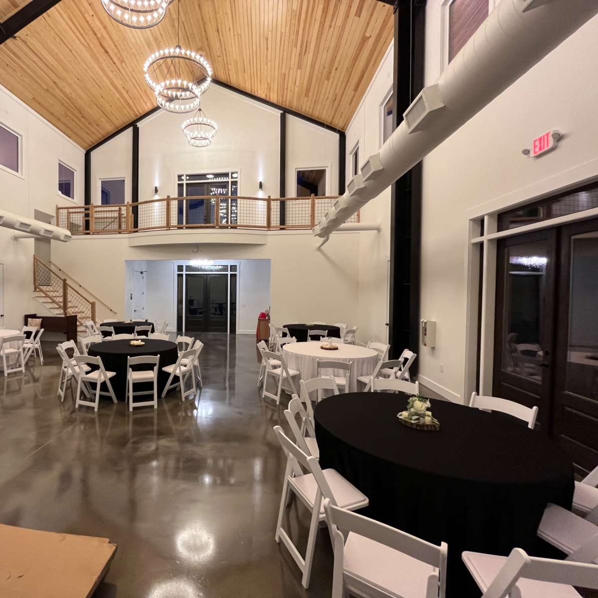 A spacious indoor venue featuring round tables with black tablecloths, white chairs, and large windows, with wooden beams and a staircase leading to an upper level.