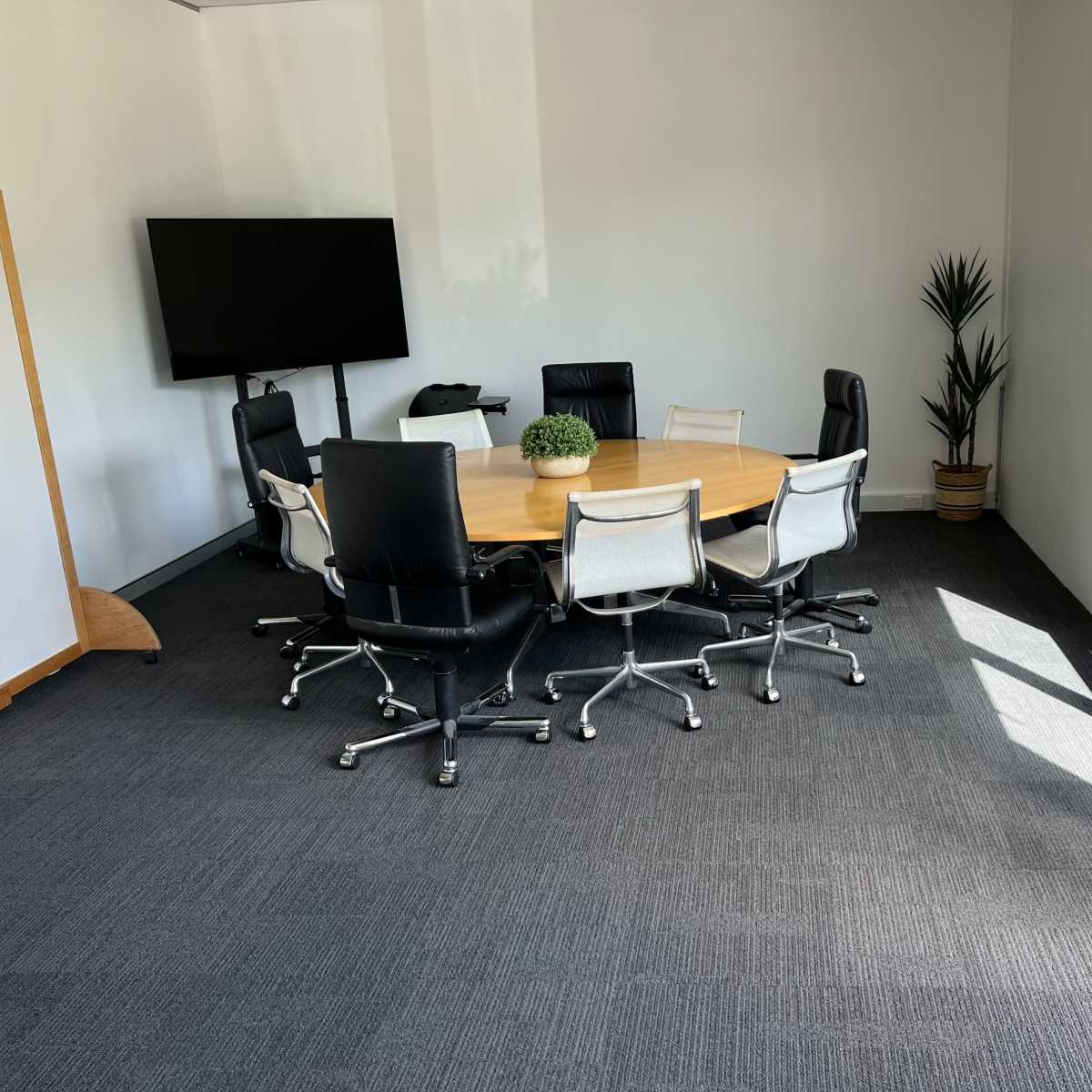 A conference room features a round wooden table surrounded by eight chairs, with a television mounted on the wall and a plant in the corner.