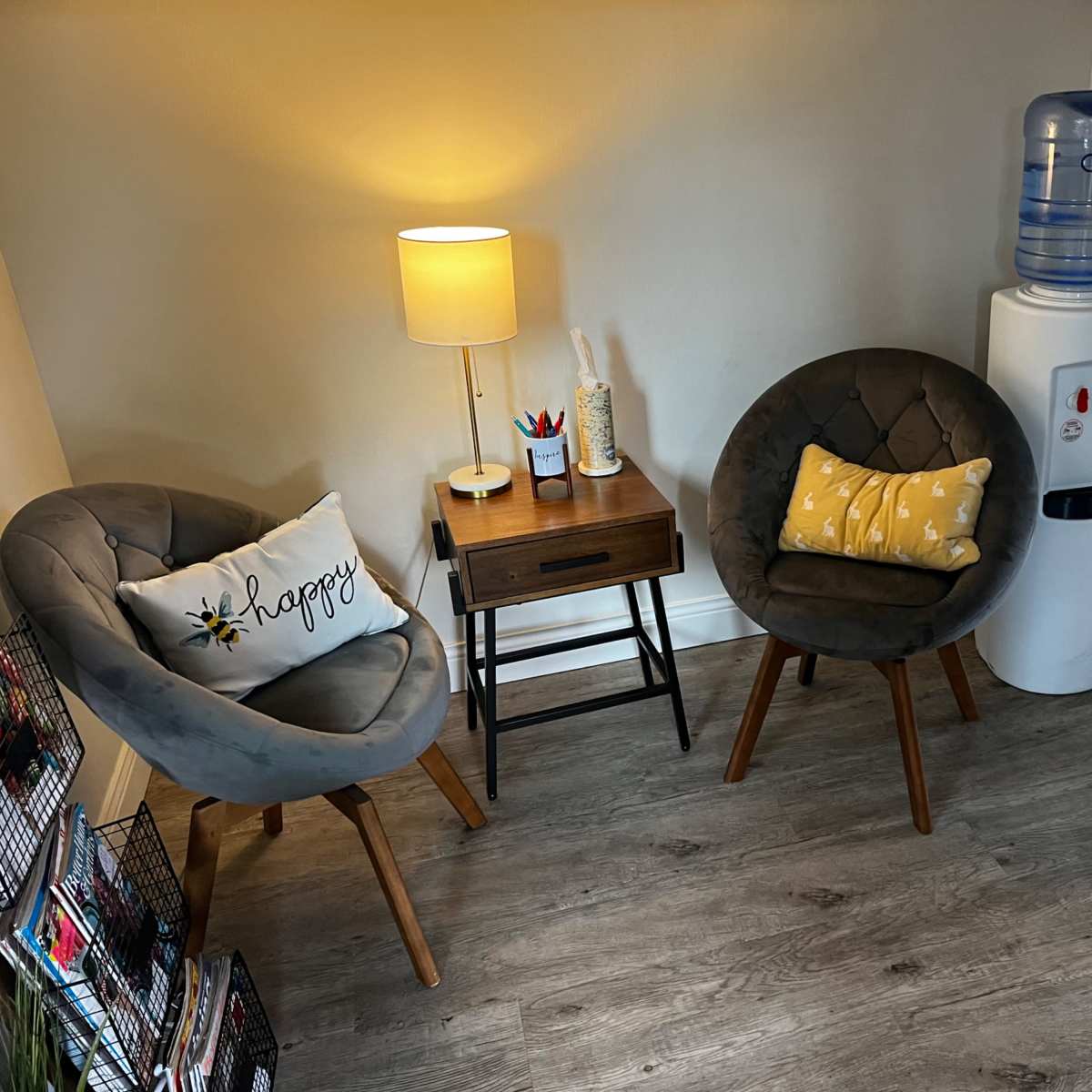 The scene features two circular chairs beside a small table with a lamp, a water cooler, and a magazine rack in a cozy corner of a room.