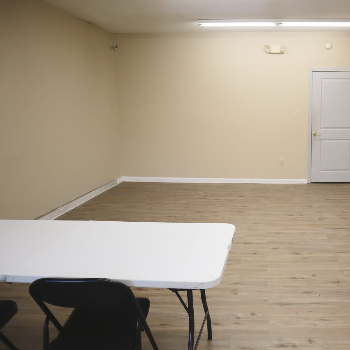 A spacious, empty room with a light brown wooden floor, a white folding table, and a closed door.