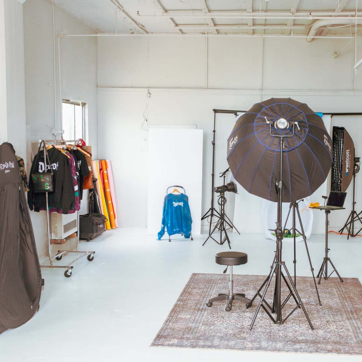 A photography studio features a lighting setup, a stool, various backdrops, and clothing racks along with a decorative rug and a plant.