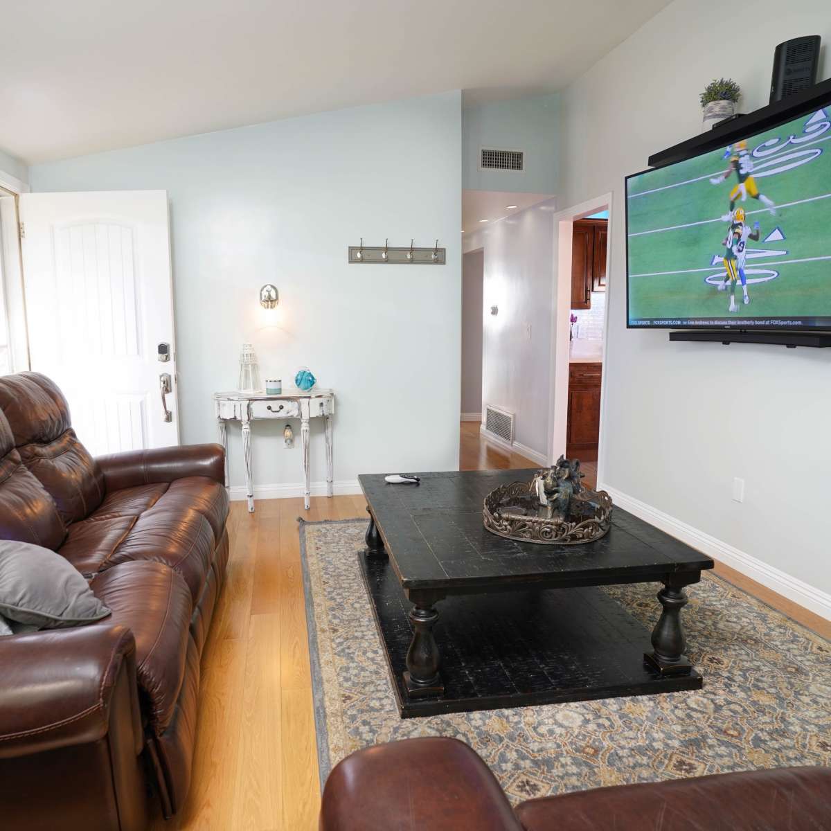 The living room features a brown leather sofa, a dark wooden coffee table, and a flat-screen television mounted on the wall, with a door visible in the background.