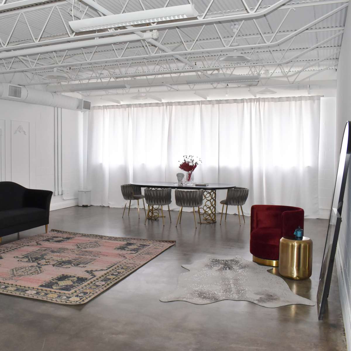 The room features a minimalist design with a black sofa, a patterned rug, a dining table surrounded by gray chairs, two red accent chairs, and large white curtains filtering light.