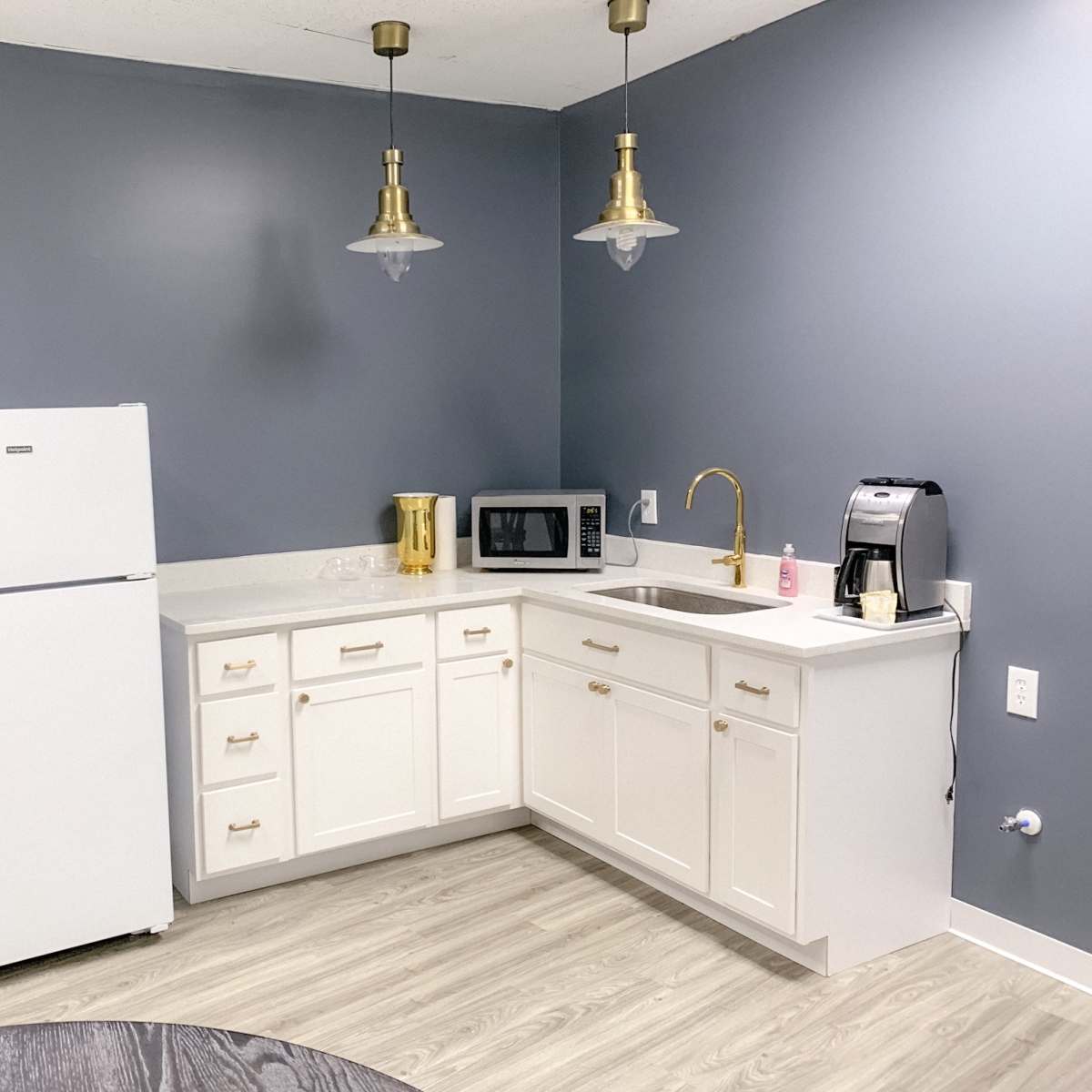 A modern kitchen with white cabinetry, a refrigerator, sink, microwave, and coffee maker against a gray wall.