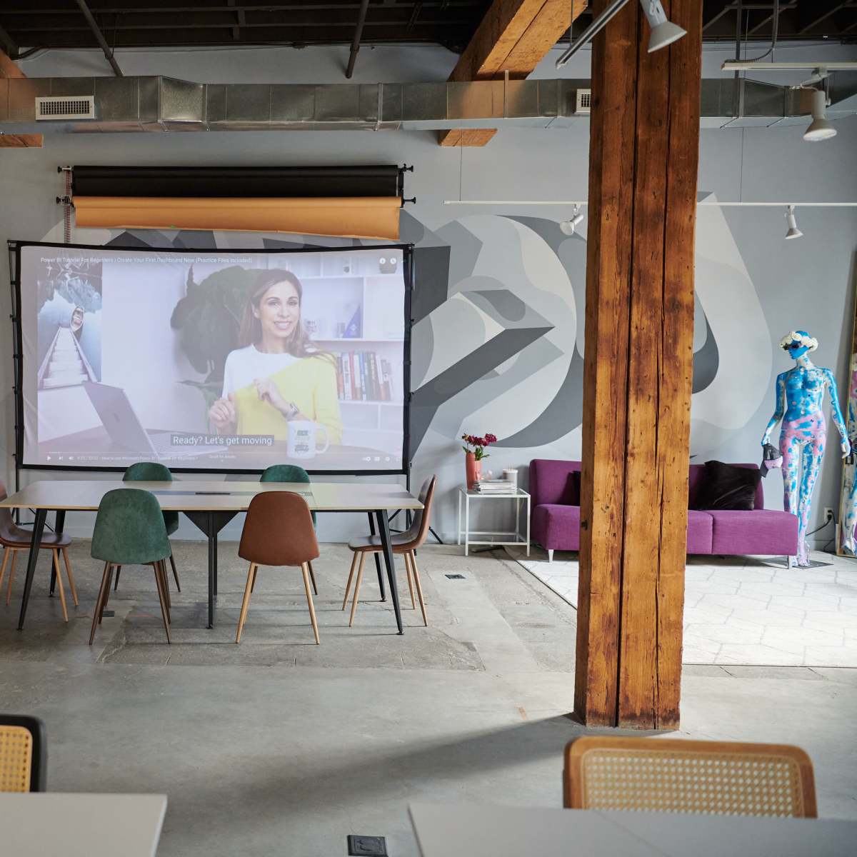 A spacious room features a large projection screen displaying a woman presenting, with a table and chairs arranged in front, and a purple sofa in the background.