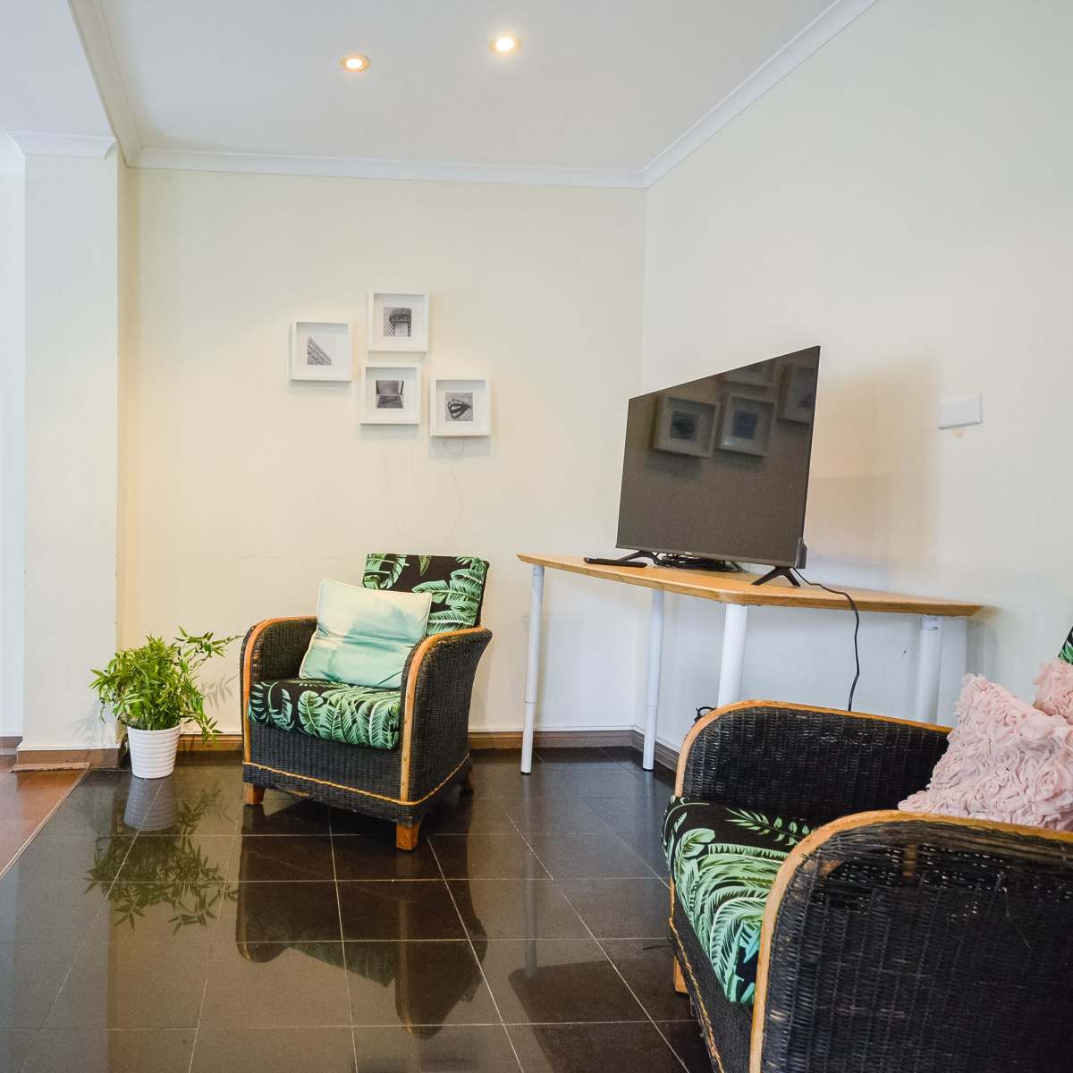 A cozy living area with a wooden table, two chairs, and a television mounted on the wall, complemented by potted plants and framed pictures.