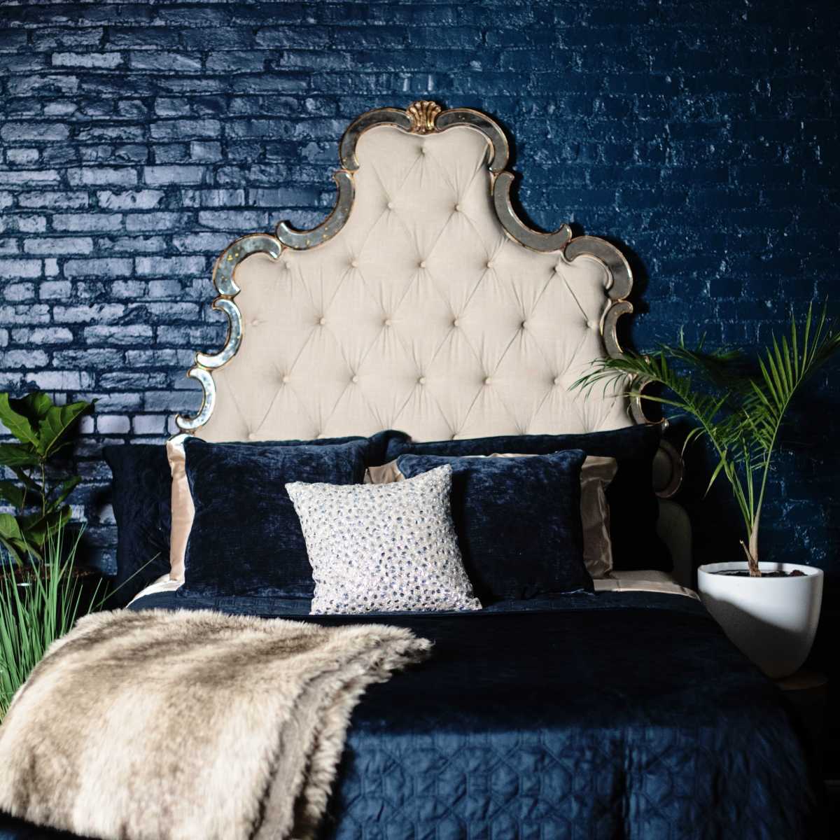 A bedroom with a large tufted headboard, navy blue walls, and a neatly made bed adorned with dark bedding and decorative pillows, accompanied by potted plants.