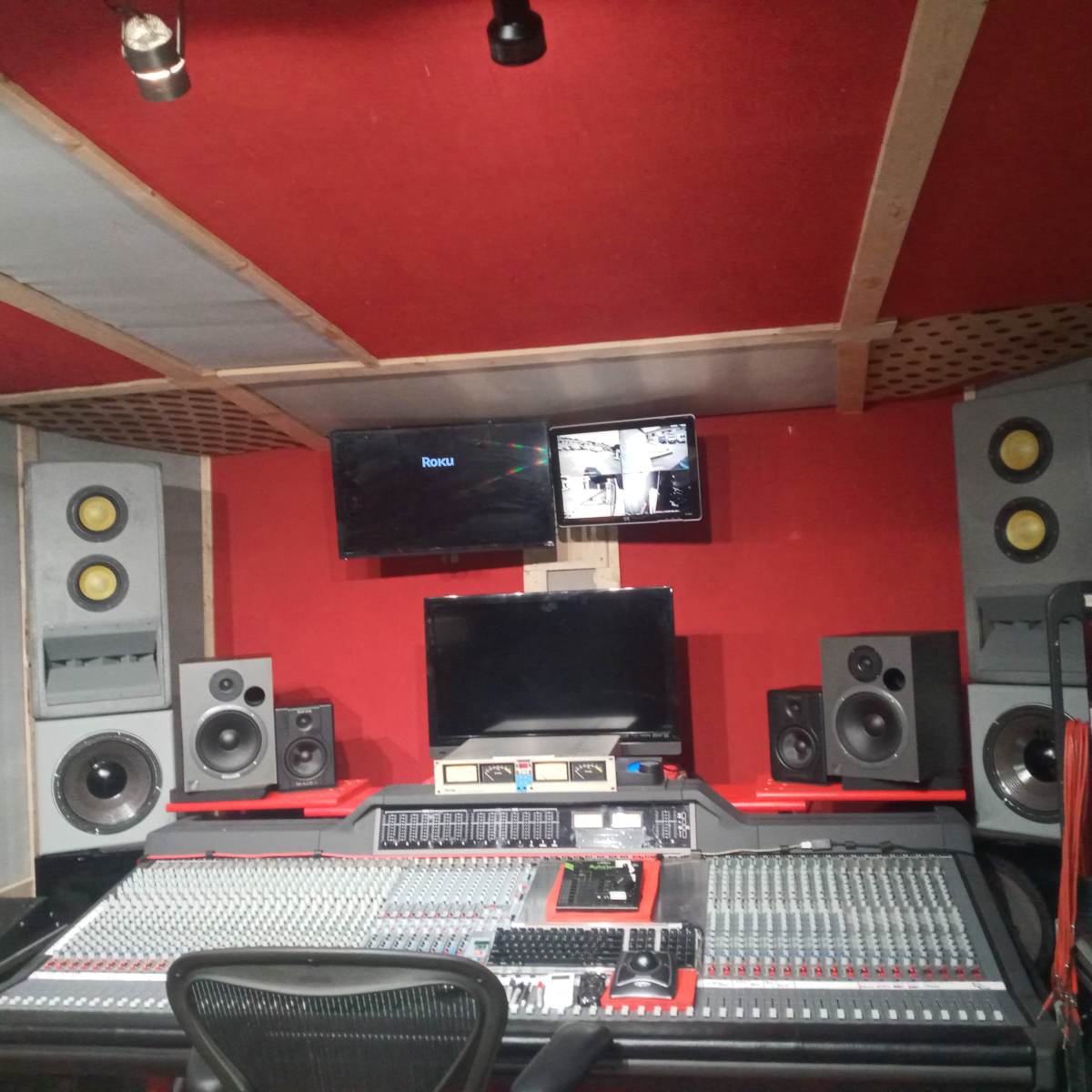 A sound recording studio featuring a large mixing console, multiple speakers, and two flat-screen monitors mounted on the walls.