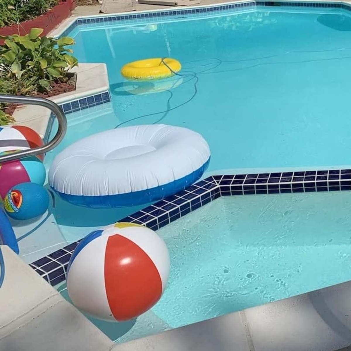 A swimming pool features colorful beach balls and inflatable floats, with a robotic cleaner in the water and steps leading into the pool.