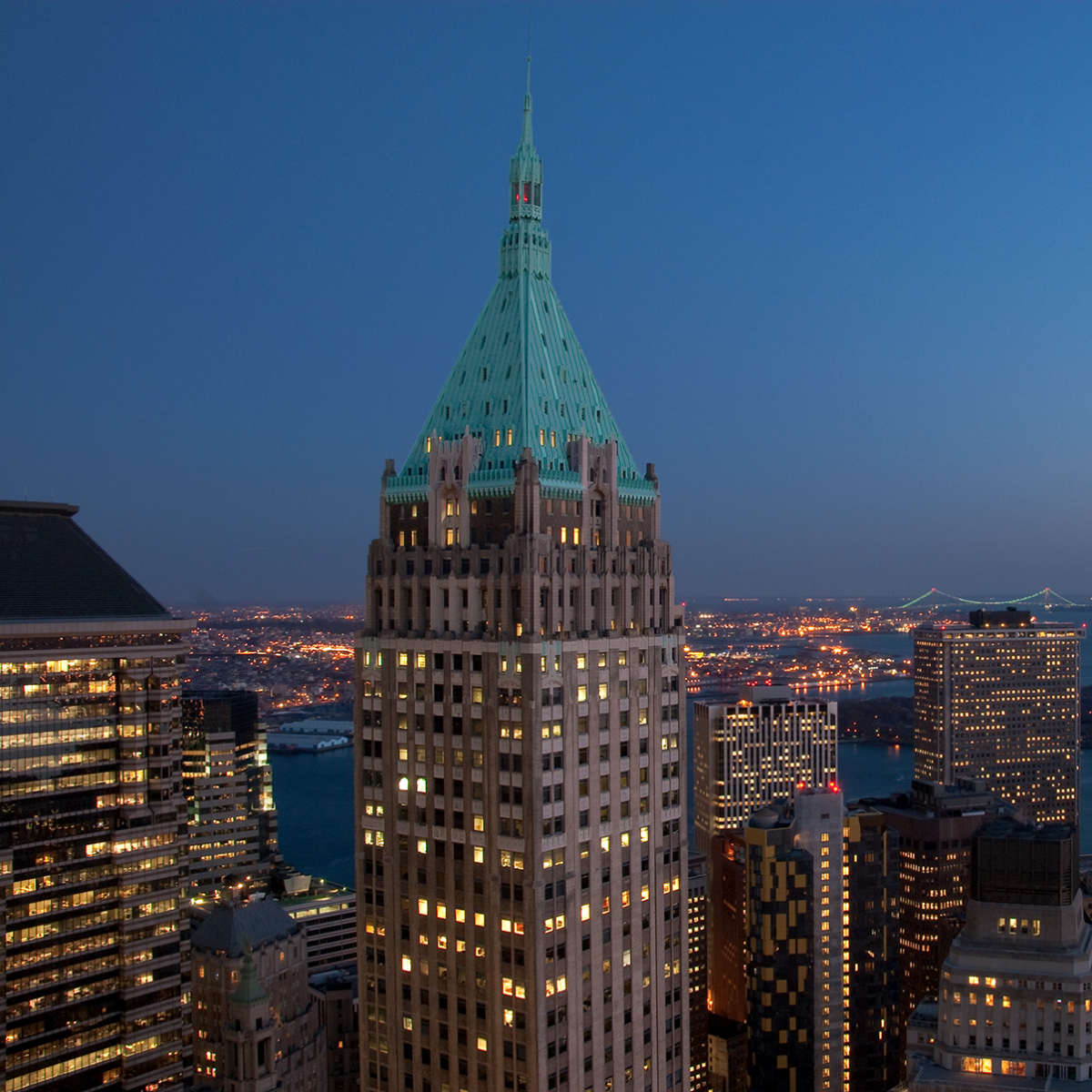 A panoramic view of a city skyline at dusk, featuring a prominent skyscraper with a green spire and a bay in the background.