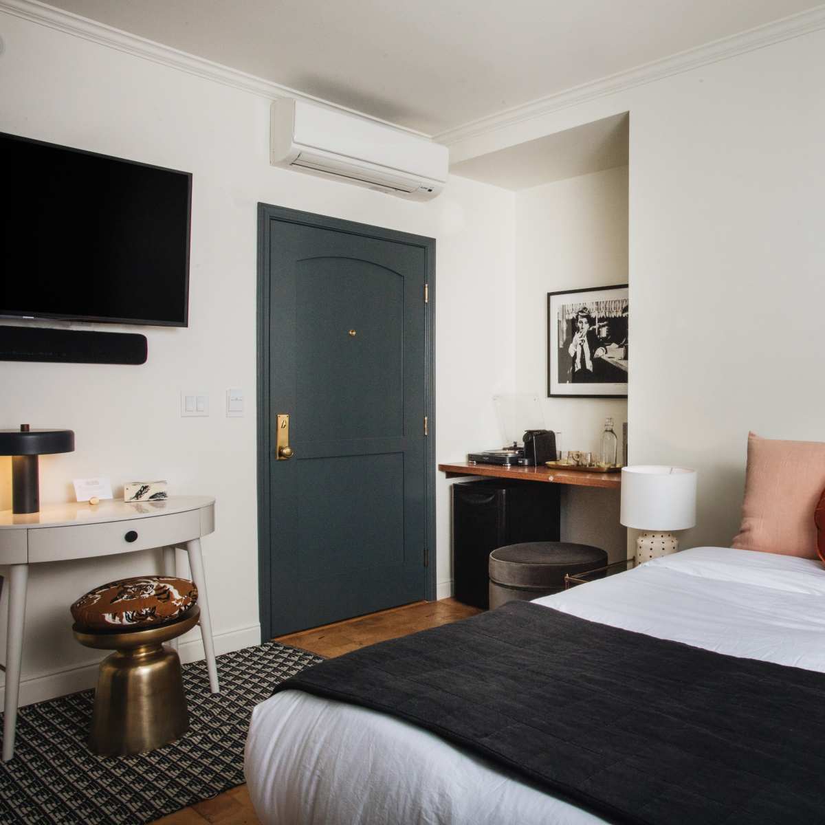 A well-furnished hotel room features a bed with decorative pillows, a bedside table, a wall-mounted television, and a small work area with a chair and cabinet.
