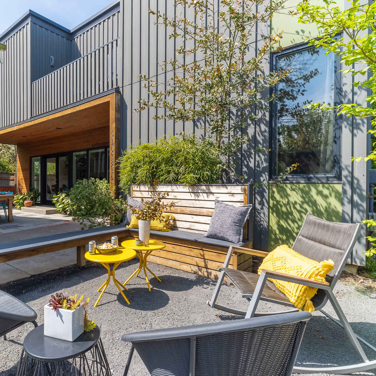 A modern outdoor seating area features a wooden bench and yellow tables surrounded by greenery and a contemporary building facade.