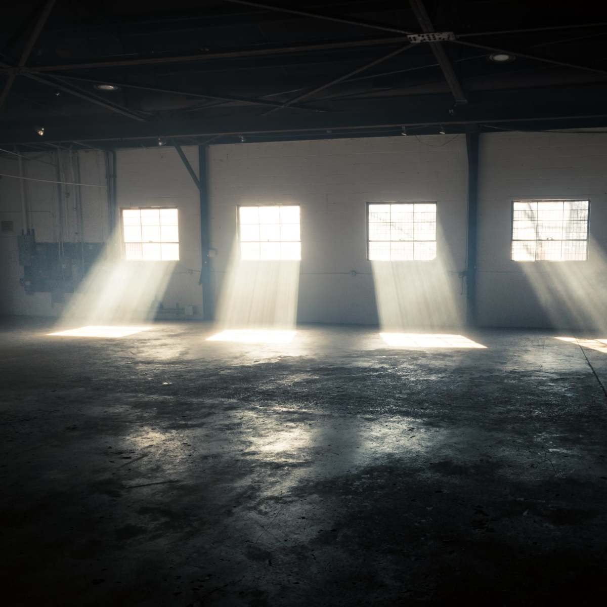 Sunlight streams through the windows of an empty industrial space, casting beams on the floor.