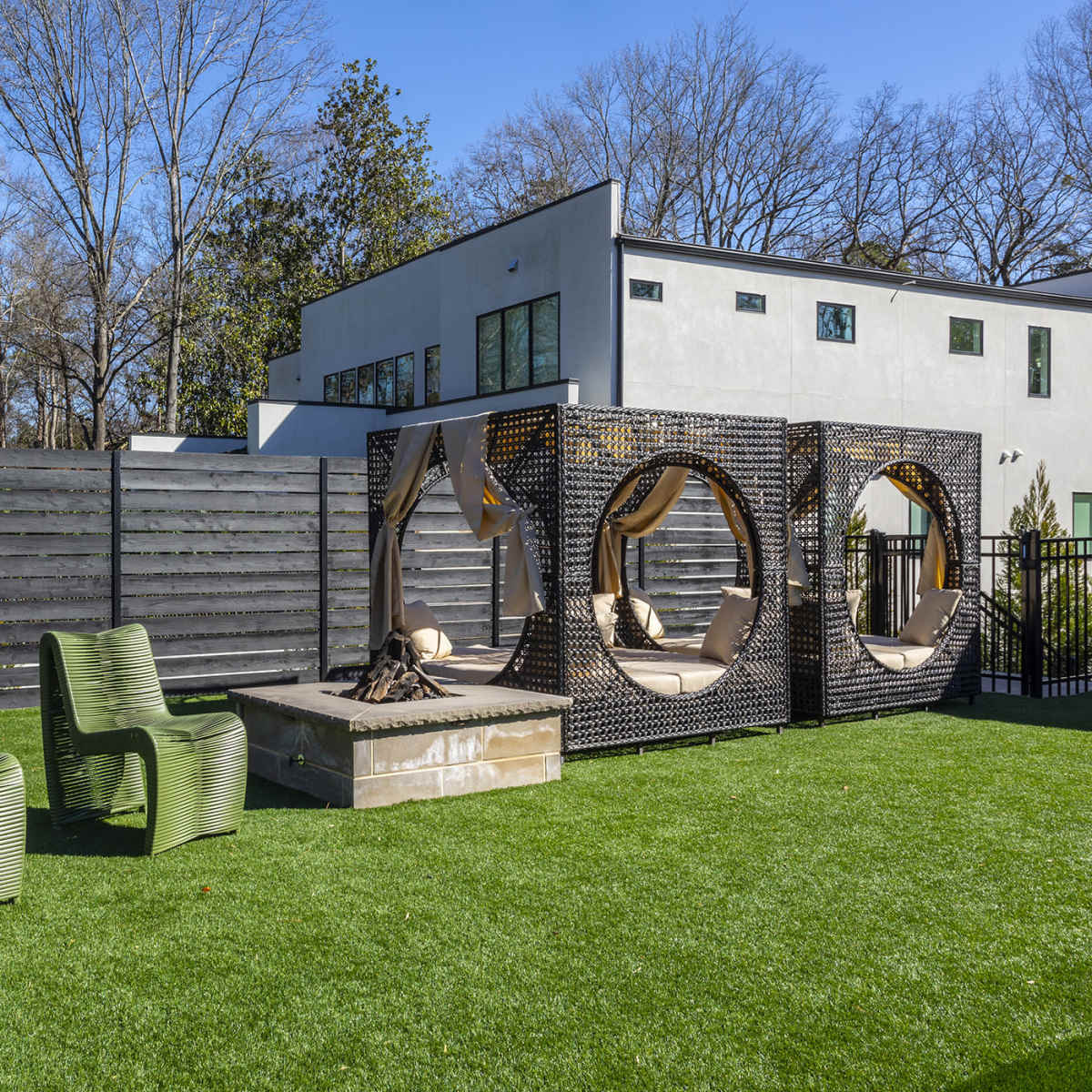 A modern outdoor space featuring two green chairs, a circular hanging chair, and a stone fire pit surrounded by faux grass and a wooden fence.