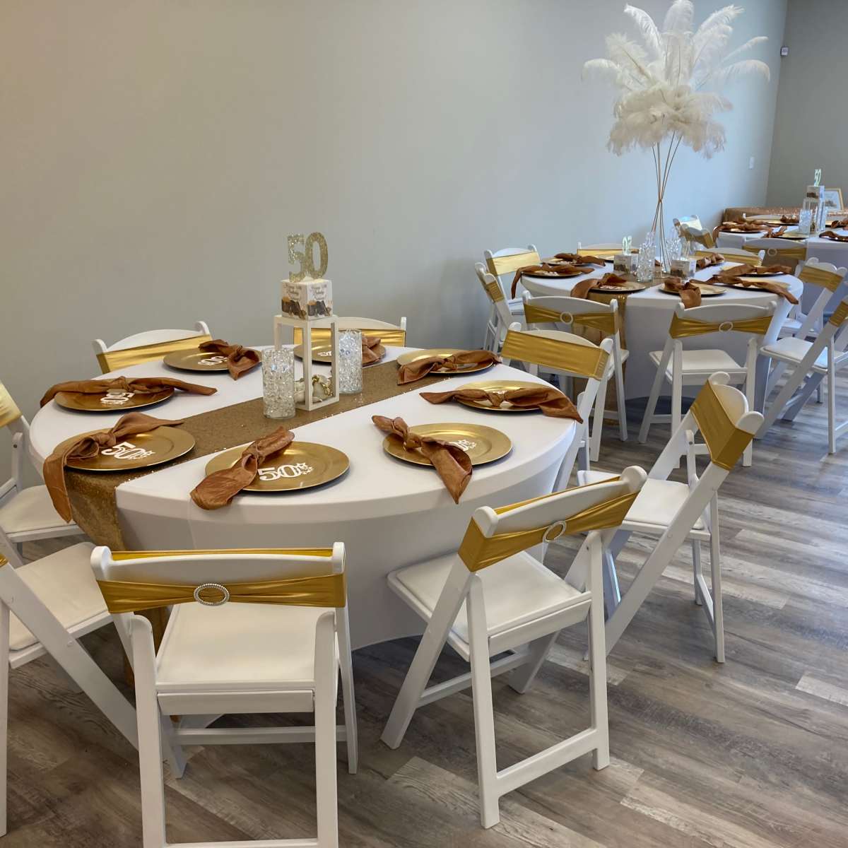 The scene shows a decorated event space with two tables set for a celebration, featuring white tablecloths, gold accents, and centerpieces.