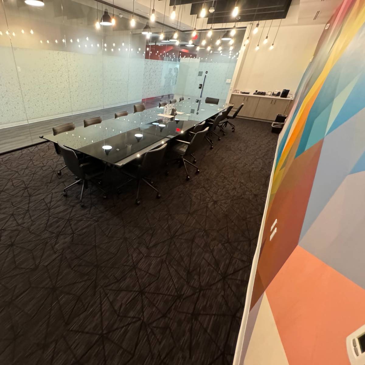 A modern conference room with a large glass table, black chairs, and a patterned carpet.