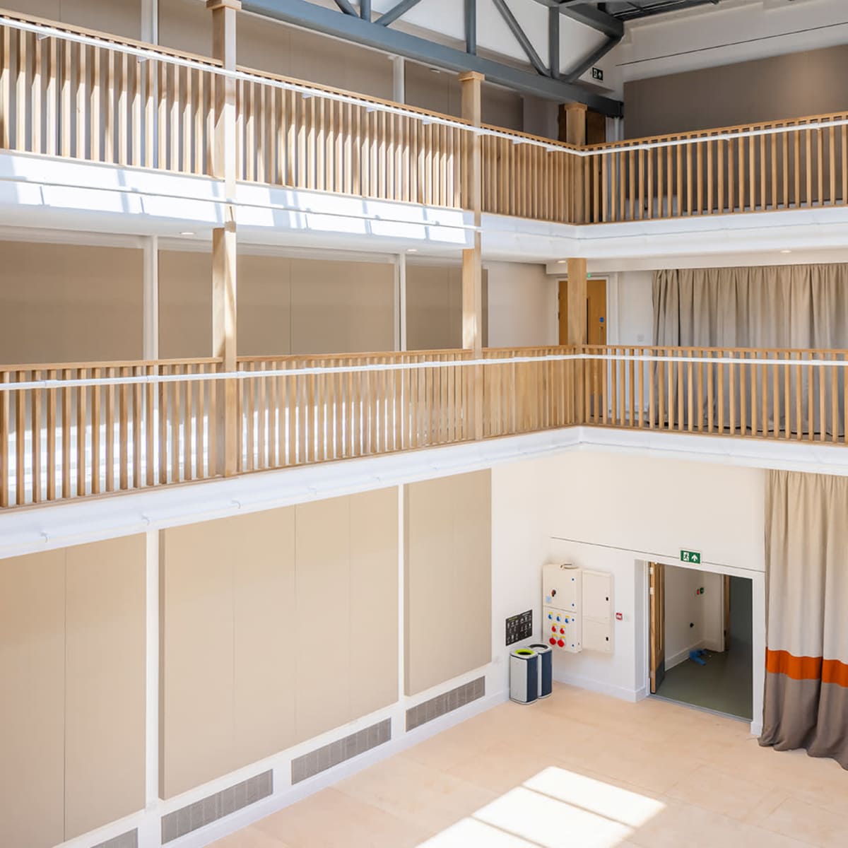 A spacious multi-level interior with wooden railings, beige walls, and large curtains, illuminated by natural light from windows above.