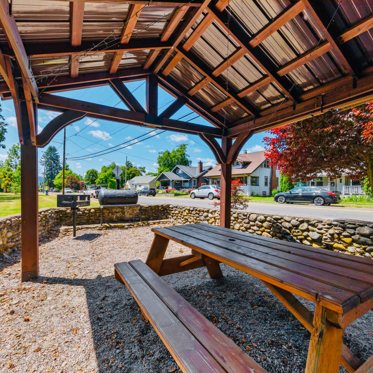A covered picnic area with a wooden table and grill is positioned next to a stone wall, overlooking a residential street lined with trees and parked cars.