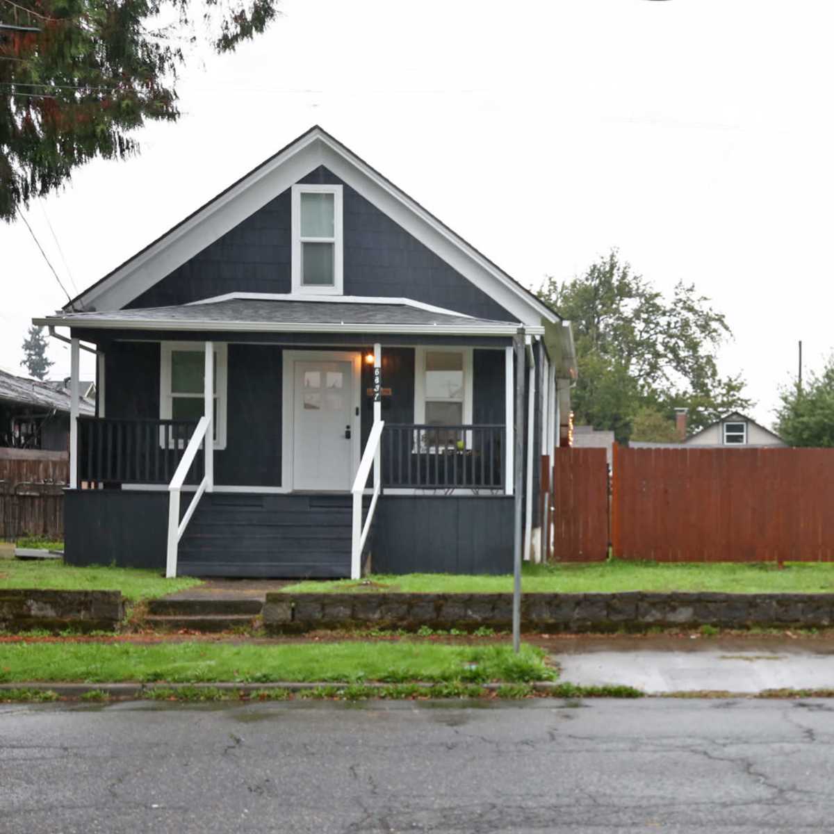 A small, dark-colored house with a front porch and a wooden fence is situated on a wet street.