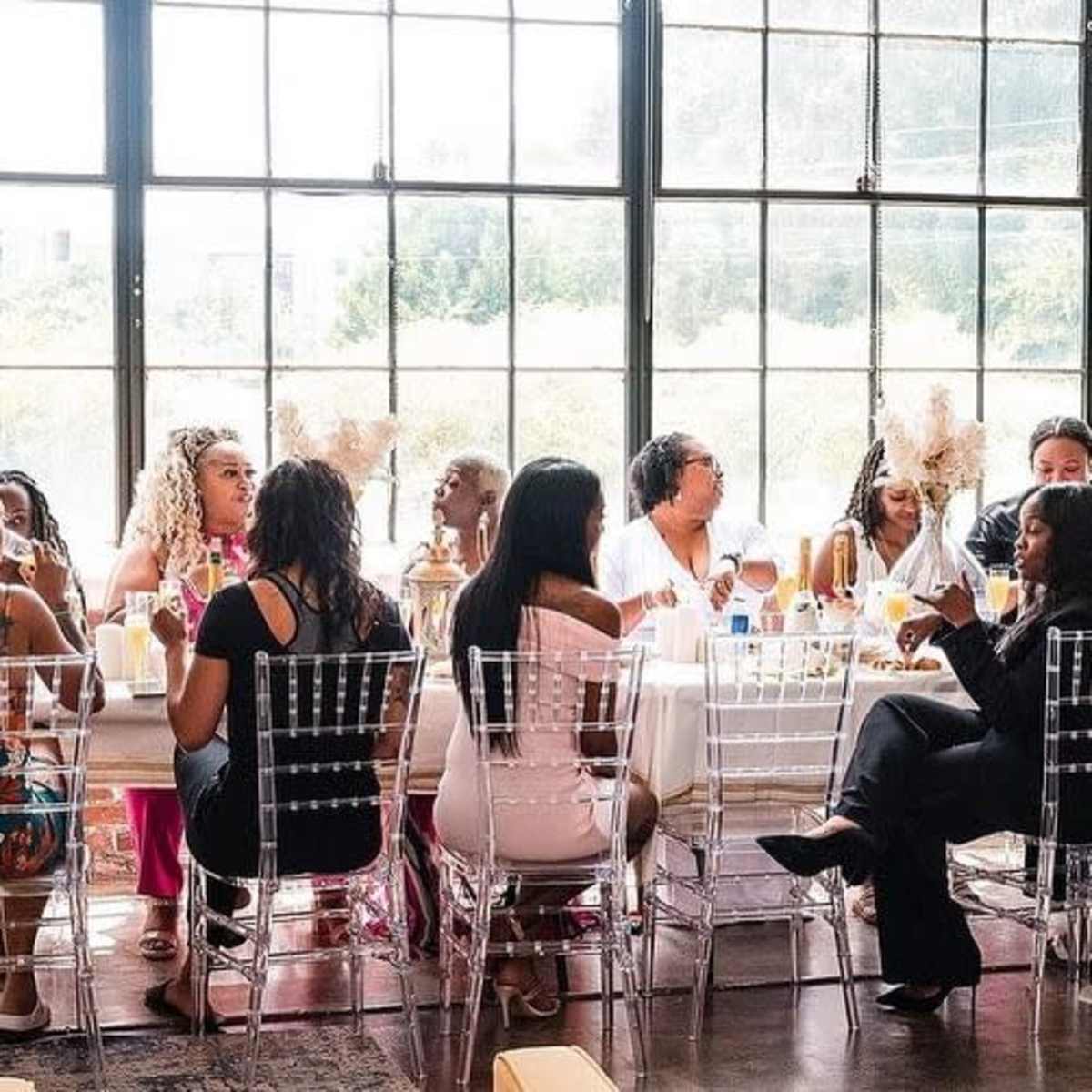 A group of people sit at a long table in a well-lit room with large windows, dining together.