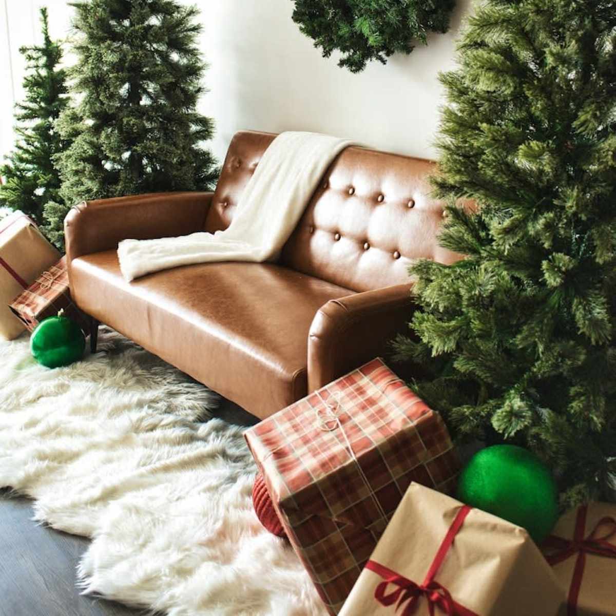 A brown leather sofa is flanked by two Christmas trees and decorated with a large wreath on the wall, surrounded by wrapped gifts.