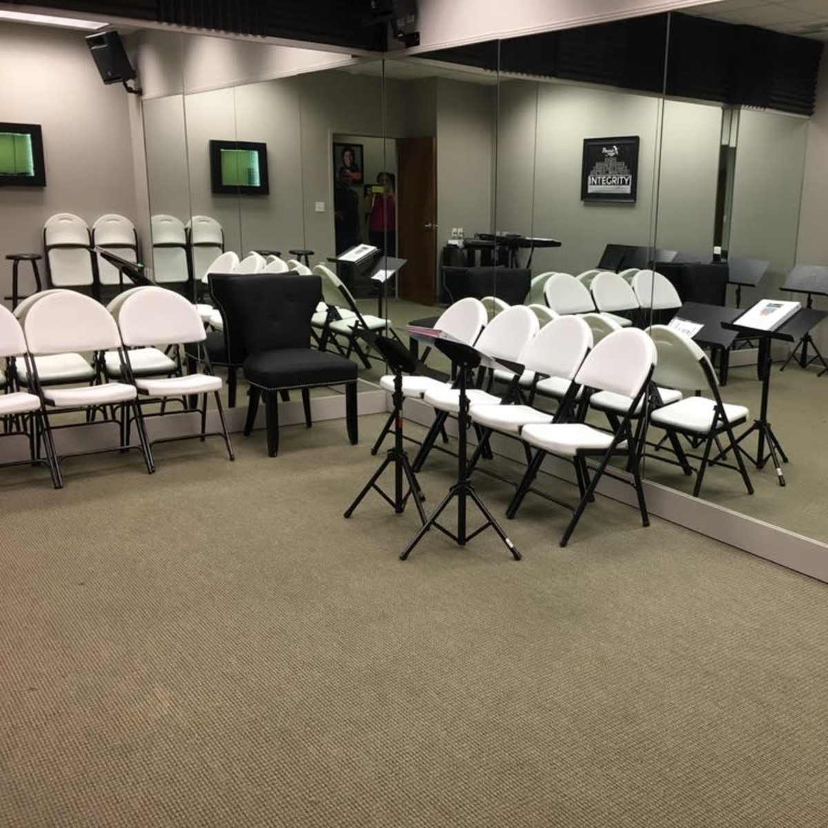 A music rehearsal room features several rows of white and black chairs, music stands, and large mirrors on the walls.