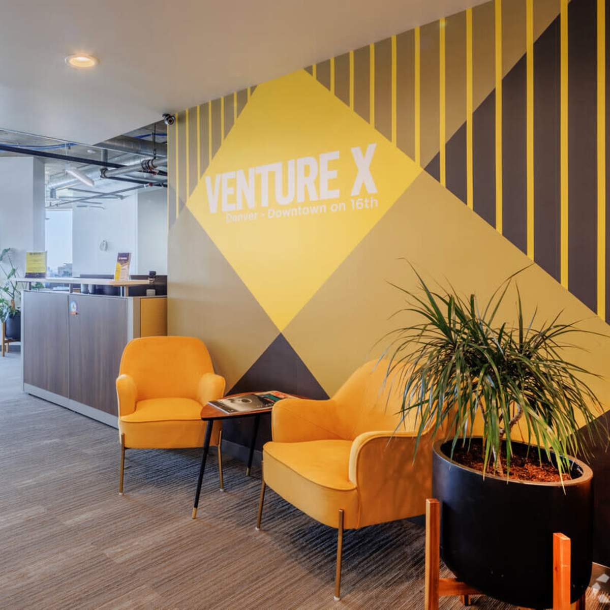 A modern office space with yellow accent chairs, a reception area, and workstations in the background under large windows.