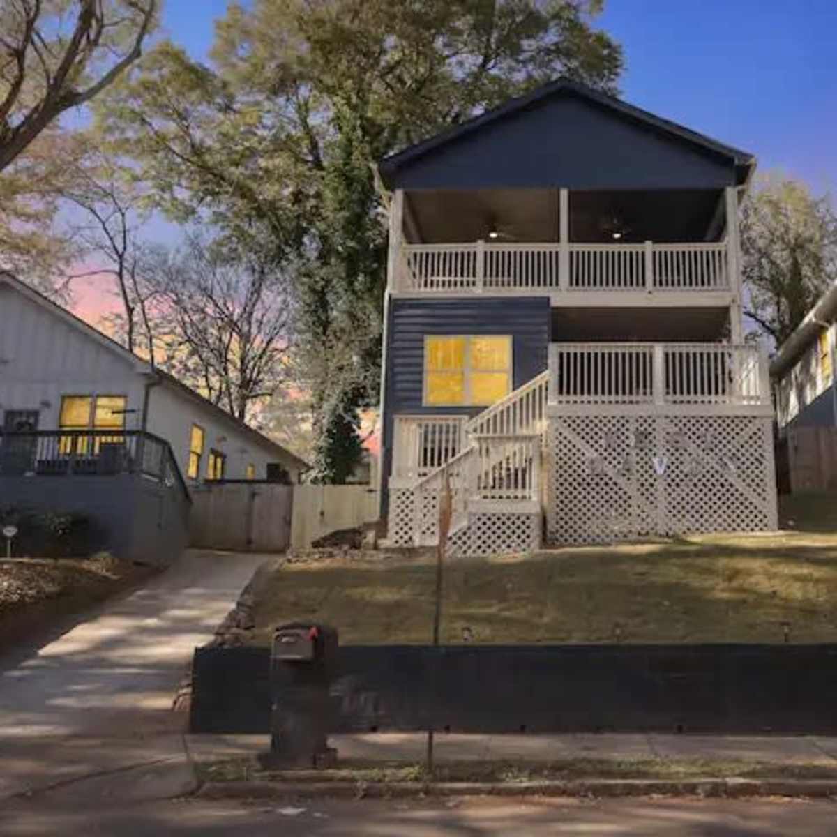 A two-story house with a balcony, located next to another house, on a street lined with trees.