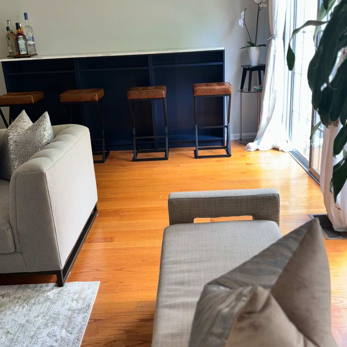 A living room with a light-colored sofa, a bar area with four stools, hardwood flooring, and large windows allowing natural light to enter.