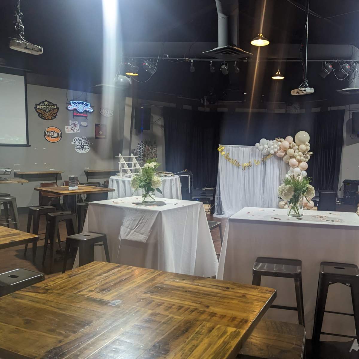 A spacious venue set up for an event, featuring tables with white tablecloths, floral centerpieces, a decorated backdrop, and screens displaying presentations.