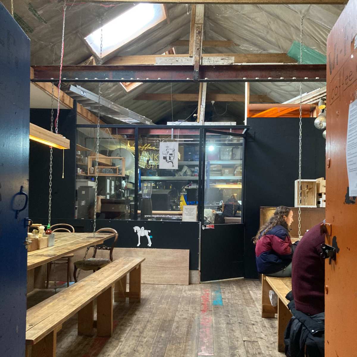 An interior space with wooden tables and benches, black walls, and a visible window at the far end.