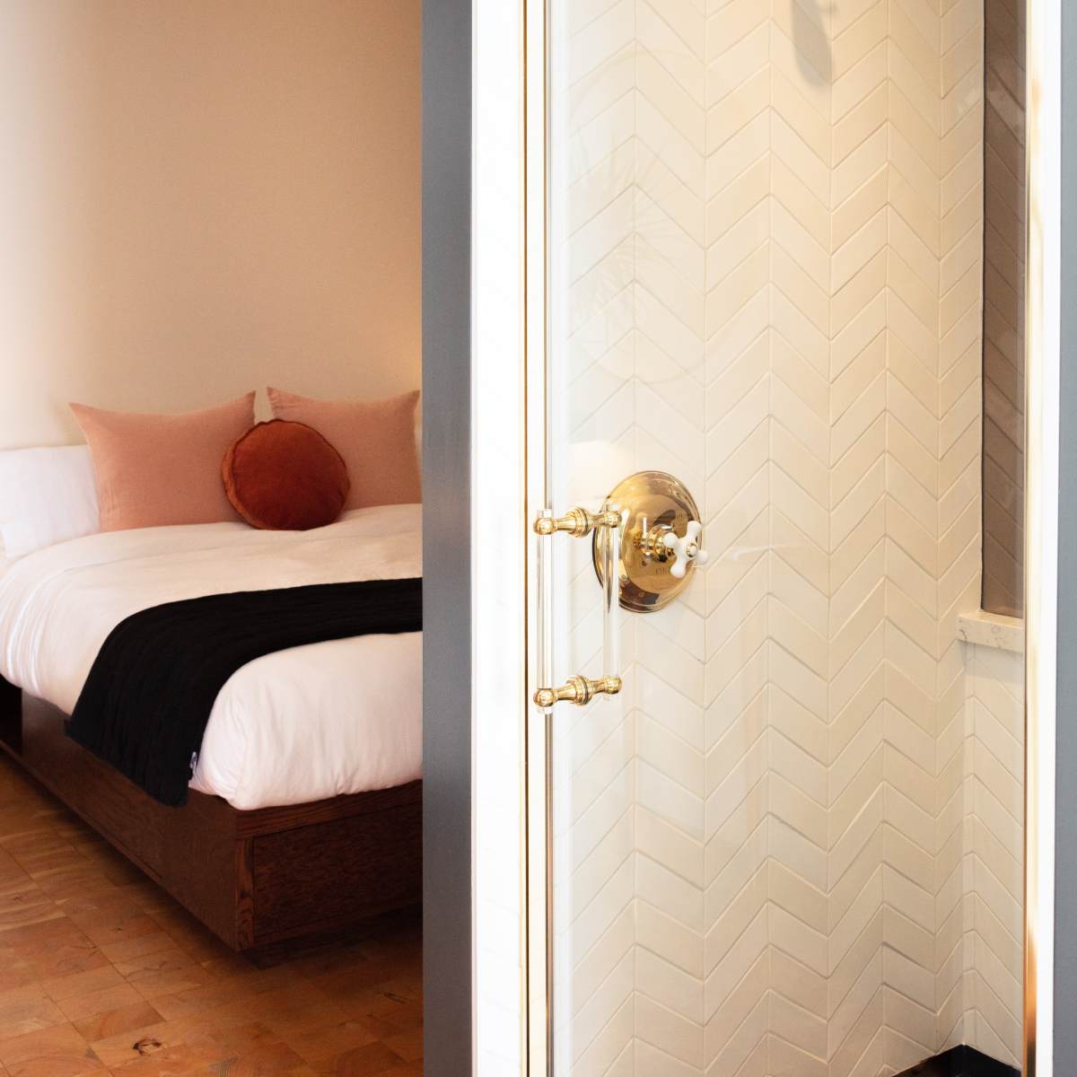 A glass shower enclosure with a gold handle stands adjacent to a bed with pink and black bedding in a well-lit room.