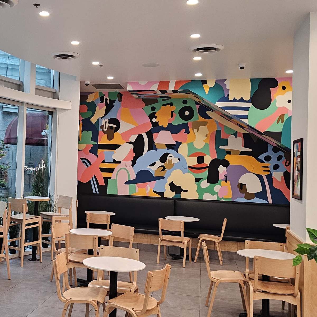 A modern cafe interior with wooden tables and chairs, large windows, and a colorful abstract mural on the wall.