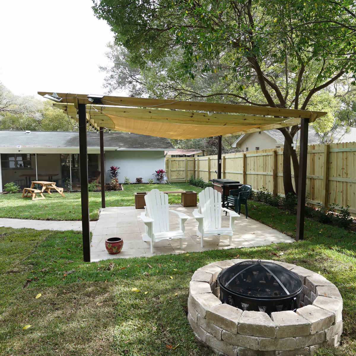 The scene features a backyard patio with a pergola, four white chairs, a grill, and a fire pit surrounded by a stone border, all set against a green lawn and wooden fence.